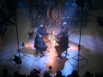 Ein Streichquartett spielt in einem dunklen Raum, umgeben von sitzendem Publikum. Stimmungsvolle Lichter beleuchten die Musiker von oben.