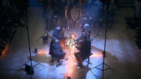 A string quartet performs in a dimly lit room, surrounded by an audience. Moody lights illuminate the musicians from above.  © Diana Lim