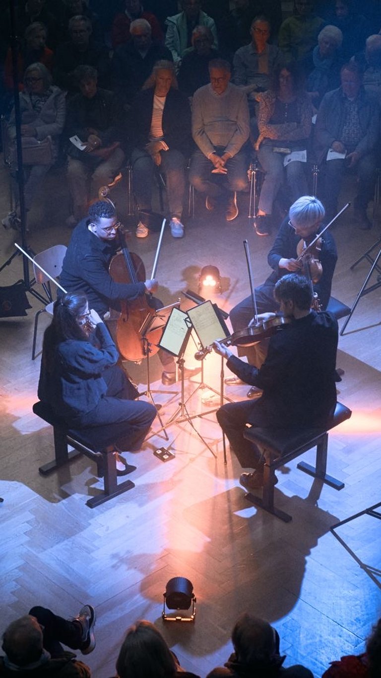 Ein Streichquartett spielt in einem dunklen Raum, umgeben von sitzendem Publikum. Stimmungsvolle Lichter beleuchten die Musiker von oben.
