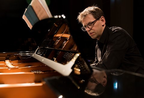 A man is intently playing the piano on stage, with open sheet music and wearing a black shirt, in dim lighting.