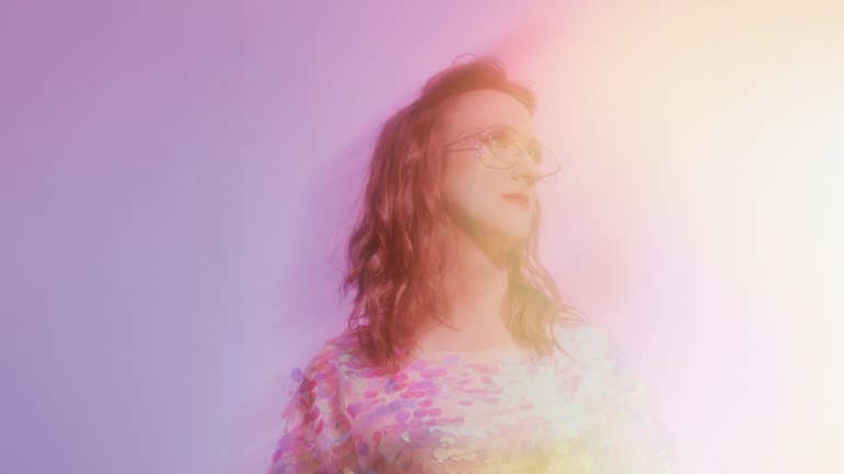 A blurred portrait of a person with long hair and glasses, enveloped in rainbow-colored light, against a purple background.