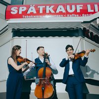 Drei Musiker:innen in formeller Kleidung stehen vor einem geschlossenen Spätkauf. Die drei halten Instrumente: Links eine Frau mit Violine; ein Mann, mittig, mit Cello; und ein weiterer Mann, rechts, hat eine Viola. Die drei scheinen gemeinsame zu spielen und blicken sich dabei an.