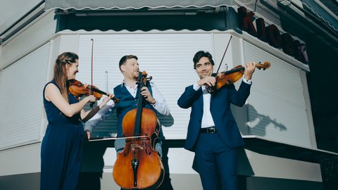 Three musicians in formal clothing are standing with each other in front of a closed shop. The three are holding instruments: On the left, a woman with a violine; a man, in the middle, with a cello; and another man, on the right, has a violo. The three are seemingly playing together and looking at each other.
