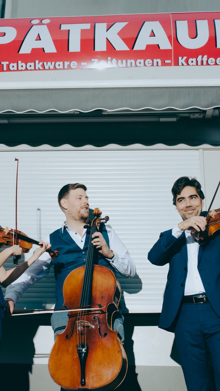 Drei Musiker:innen in formeller Kleidung stehen vor einem geschlossenen Spätkauf. Die drei halten Instrumente: Links eine Frau mit Violine; ein Mann, mittig, mit Cello; und ein weiterer Mann, rechts, hat eine Viola. Die drei scheinen gemeinsame zu spielen und blicken sich dabei an.