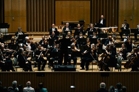 Ein Orchester spielt in einem Konzertsaal. Der Dirigent steht in der Mitte, umgeben von Musikern mit verschiedenen Instrumenten.