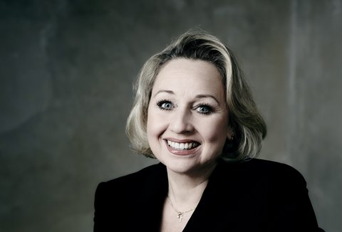 Eine Frau im schwarzen Anzug posiert für die Kamera. Ihre Haare sind blond und sie lächelt.