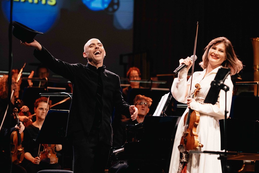 Ein Dirigent und eine Geigerin stehen freudig auf einer Bühne vor einem Orchester, während eines musikalischen Auftritts.