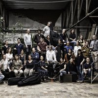 A large group of people poses together in an industrial space. Some hold musical instruments, others stand casually.