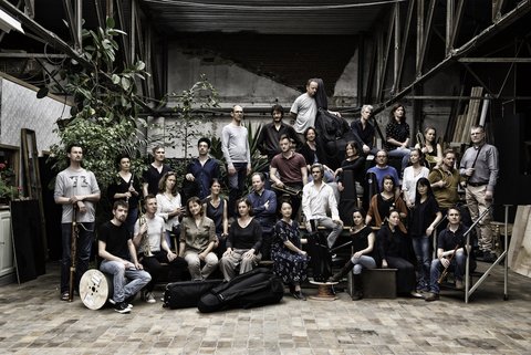 Eine große Gruppe von Menschen posiert gemeinsam in einem industriellen Raum. Einige halten Musikinstrumente, andere stehen lässig.