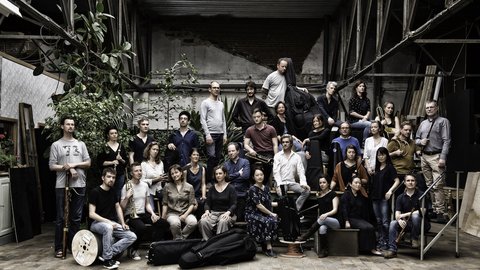 Eine große Gruppe von Menschen posiert gemeinsam in einem industriellen Raum. Einige halten Musikinstrumente, andere stehen lässig.