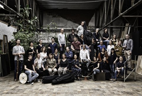 A large group of people poses together in an industrial space. Some hold musical instruments, others stand casually.
