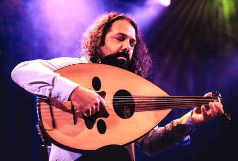 Ein Mann spielt Oud, die arabische Laute. Er hält das Instrument vor der Brust und zupft. Seine Augen sind geschlossen. Im Hintergrund ist Bühnenscheinwerferlicht und Neben zu sehen.