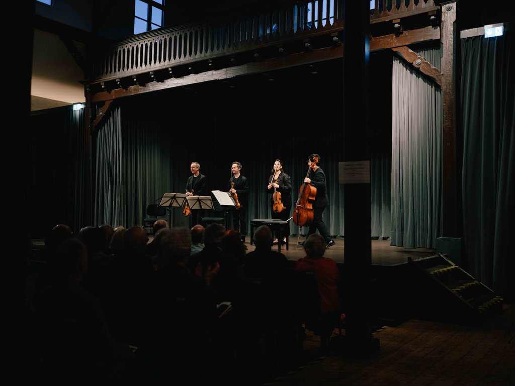 A string quartet performs on a stage with green curtains, illuminated by warm light. The audience sits in a dimmed room.