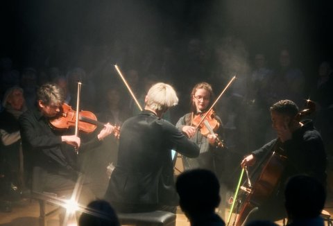 A string quartet performs in a dimly lit room. The audience sits in a semicircle, attentively listening to the concert.